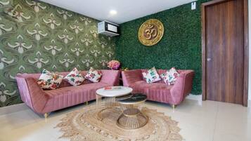 Une pièce avec un papier peint à motifs verts, un canapé rose agrémenté de coussins à fleurs, une table basse ronde et un climatiseur fixé au mur.
