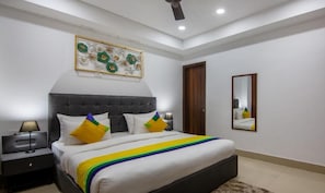 A bedroom with a large bed, a nightstand, a lamp, a ceiling fan, a wall-mounted mirror, and a decorative wall piece. - Hotel Divine India Noida Sector 135 (Noida)