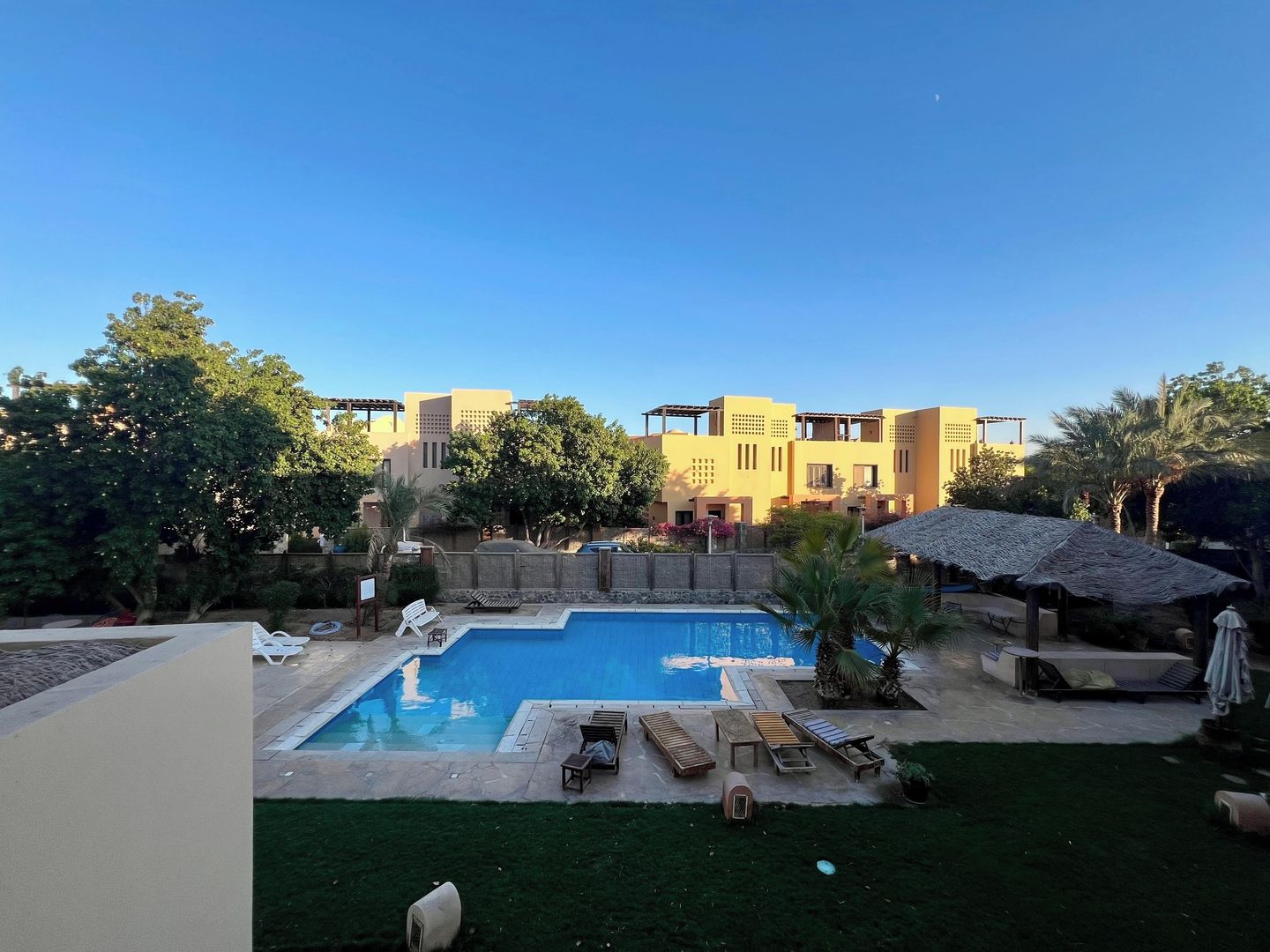 Ein Pool mit Liegestühlen und einer strohgedeckten Struktur, umgeben von Grünflächen und Gebäuden.