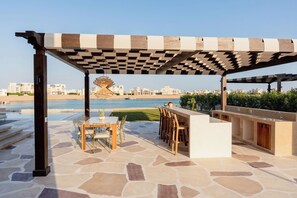 An outdoor patio with a pergola, a dining table with chairs, a bar area, and a view of the sea and buildings in the background.