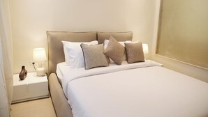 Una cama bien hecha con ropa blanca y varias almohadas, una lámpara de noche y un florero en la mesita de noche.