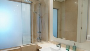 Moderni kylpyhuone, jossa on lasiseinällä varustettu suihku, valkoinen pesuallas ja suuri peili.