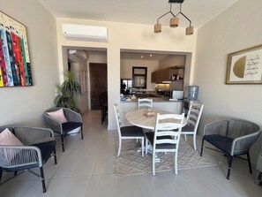 A dining area with a round table and chairs, a kitchen in the background, and a potted plant.