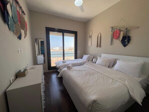 Een hotelkamer met twee bedden, een bureau, een stoel en uitzicht op de stad door een groot raam.