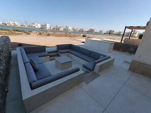 A modern outdoor seating area with a sectional sofa and a coffee table, overlooking a beach and cityscape.