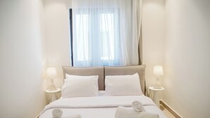 Een hotelkamer met een bed, twee bedlampen, een raam met gordijnen en twee opgerolde handdoeken op het bed.