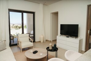 Una sala de estar moderna con un sofá, sillones, una mesa de centro, un televisor sobre un mueble blanco y un gran ventanal corredizo que da a un área exterior.