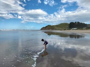 A beach with a person standing in shallow water, a clear sky, and distant hills. - Classic Kiwi bach escape with cliff views (Kairākau)