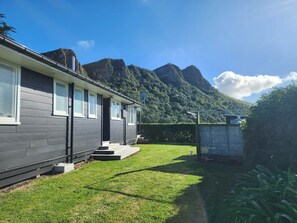 A modern two-story house with a well-maintained lawn and a mountain backdrop. - Classic Kiwi bach escape with cliff views (Kairākau)