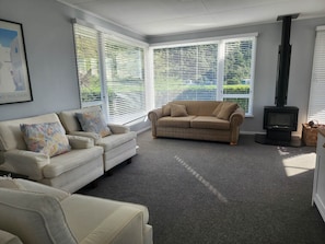 A living room with a sofa, a smaller couch, a fireplace, and large windows with blinds. - Classic Kiwi bach escape with cliff views (Kairākau)
