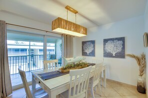 Un comedor con una mesa de vidrio, sillas blancas, una lámpara colgante y dos cuadros de árboles en la pared.