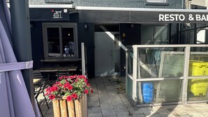 Un bâtiment sur lequel est apposée une enseigne indiquant « RESTAURANT ET BAR », et devant lequel se trouve un bac à fleurs en bois orné de fleurs rouges.