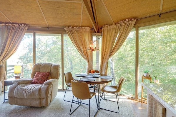 A room with a round table, chairs, a sofa, and a view of trees through large windows.