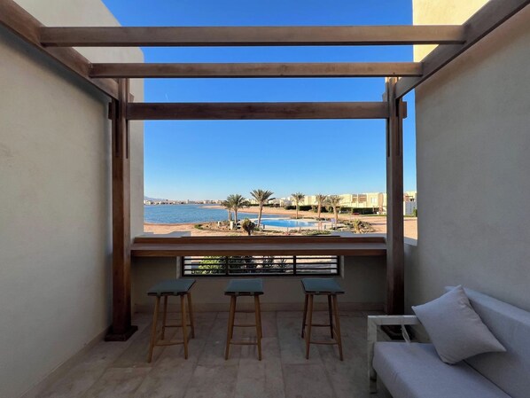 A balcony with a view of a pool and beach, wooden stools, and a white sofa.