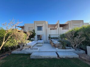 A modern two-story building with a flat roof, a covered outdoor seating area, and a well-maintained garden with various plants and a lawn.