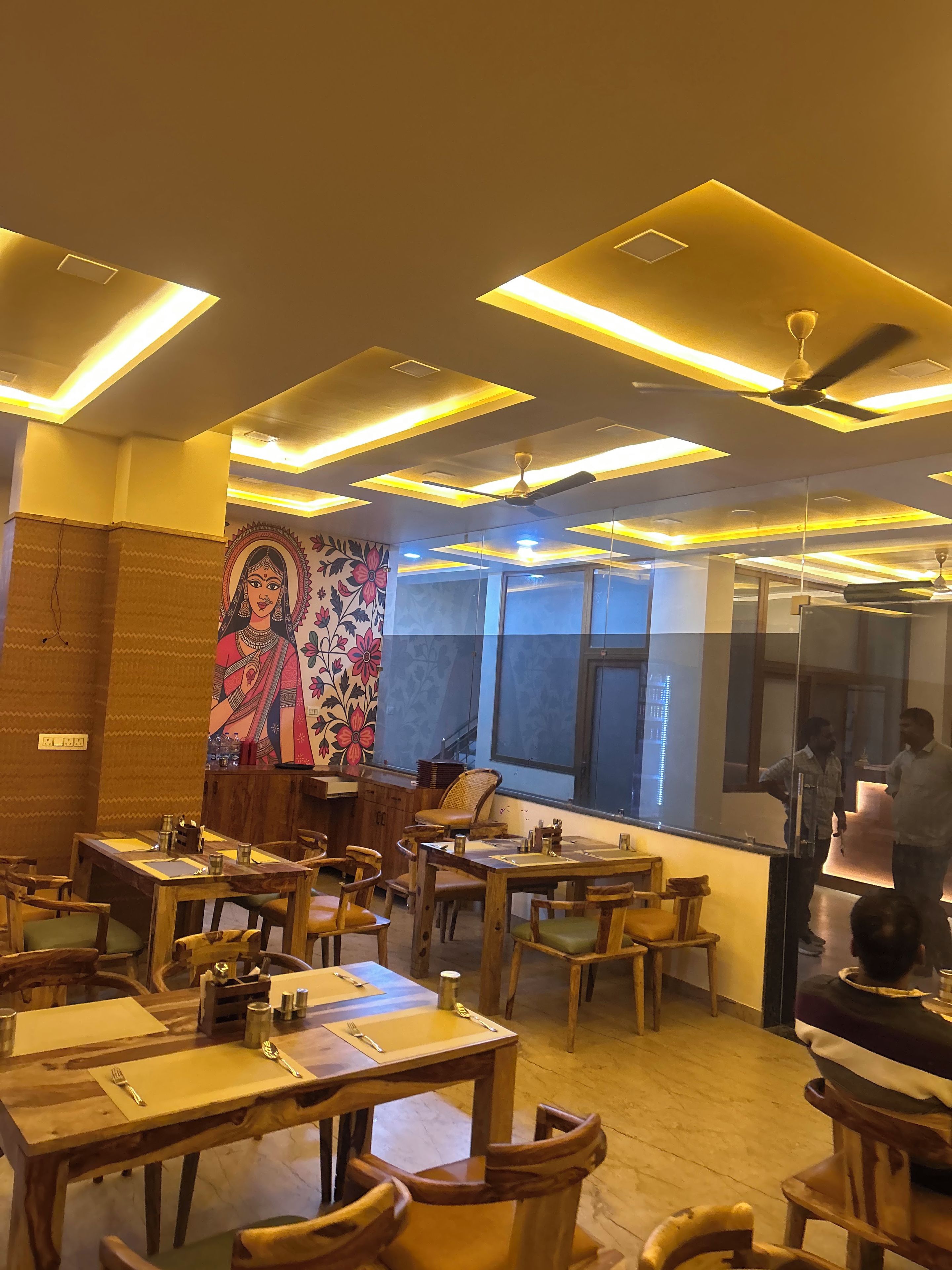 Interior de restaurante com mesas e cadeiras de madeira, um mural na parede e teto com iluminação embutida.