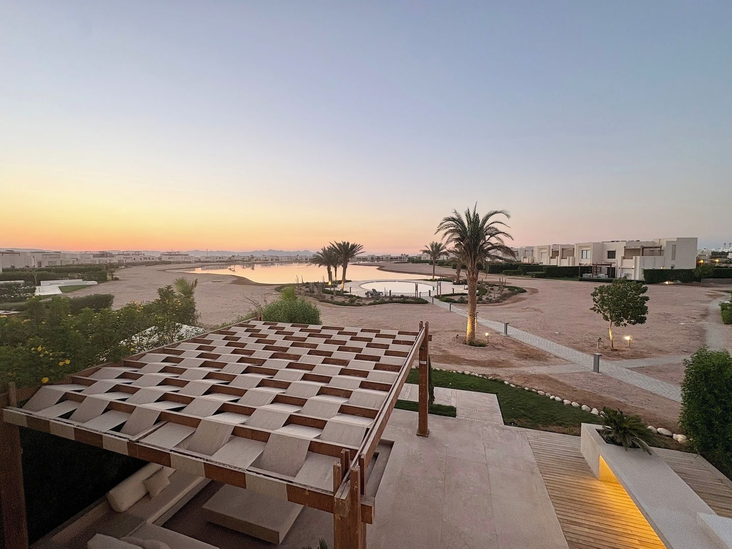 A modern outdoor area with a wooden pergola, palm trees, and a view of a body of water and buildings in the distance.