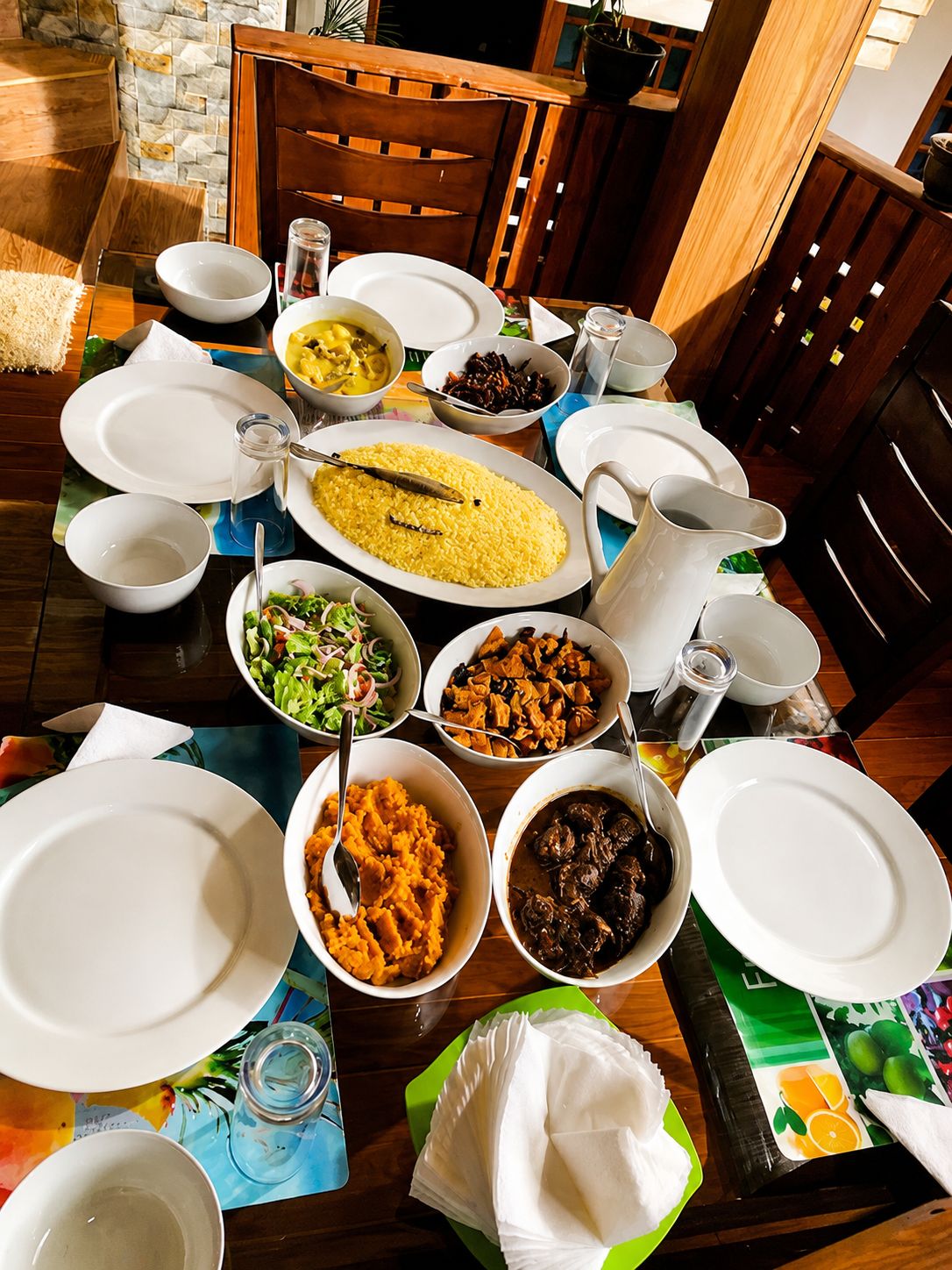 A dining table set with various dishes, including rice, salad, and meat, in a room with wooden furniture and a stone wall.
