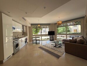 A modern living room with a sofa, a coffee table, and a TV. There is a kitchen area with a washing machine and cabinets. Large windows provide natural light and views of the outside. - Cozy 1BR GF Mangroovy Residence (Hurghada)
