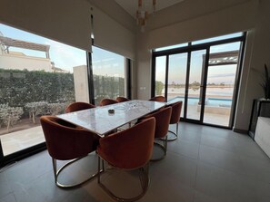 A modern dining area with a marble table and orange chairs, large windows with a view of a pool and greenery. - Luxury 4BR Cyan Villa • Private Pool (Hurghada 2)