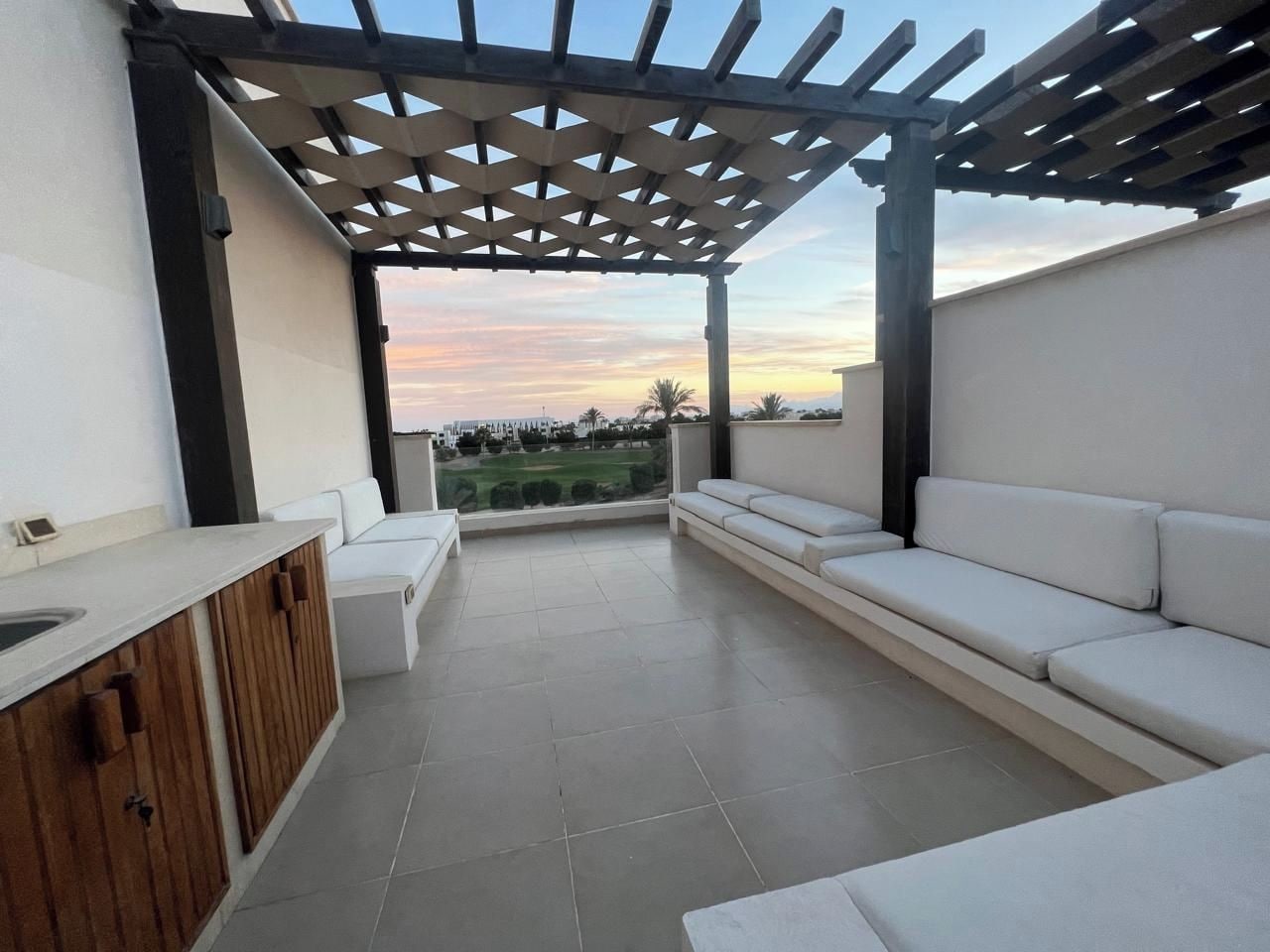 Un patio exterior con pérgola, sofás blancos, un mueble de madera y vistas del paisaje.