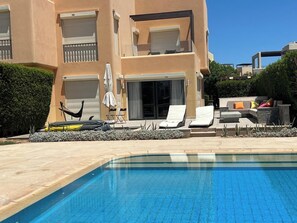 A swimming pool with lounge chairs and a hammock, surrounded by a building with balconies and a hedge. - Tawila 3BR Family Townhouse • Lagoon Retreat Villa (Hurghada 2)