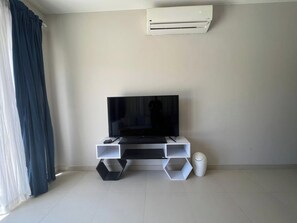 Una sala de estar con un televisor de pantalla plana sobre un mueble blanco, un cubo de basura blanco y un aire acondicionado instalado en la pared.