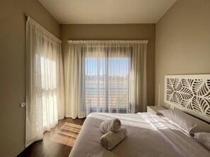 Habitación de hotel con una cama grande, cabecera con un panel decorativo y vistas a una piscina a través de cortinas translúcidas.