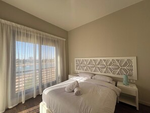 A hotel room with a large bed, a nightstand, a decorative headboard, and a view through sheer curtains.