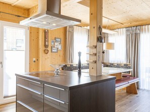 A modern kitchen with a central island, wooden beams, and a large window with curtains. - One-bedroom Cabin (Sankt Georgen am Kreischberg)