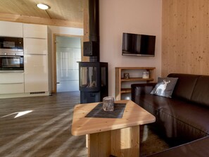 A cozy living area with a wooden table, a leather sofa, a TV mounted on the wall, and a kitchen in the background. - Cabin With 1 Bedroom (Sankt Georgen am Kreischberg)