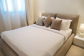 Una cama bien hecha con cabecera, lámpara de noche y ventana con cortinas traslúcidas.