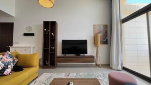 A living room with a sofa, a television, a bookshelf, and a painting on the wall. - Stylish G-Cribs 2BR • Pool View (EL Gouna)