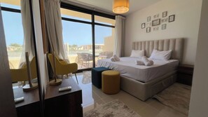 A hotel room with a bed, a desk with a remote control, a chair, and a view of the outside. - Stylish G-Cribs 2BR • Pool View (EL Gouna)