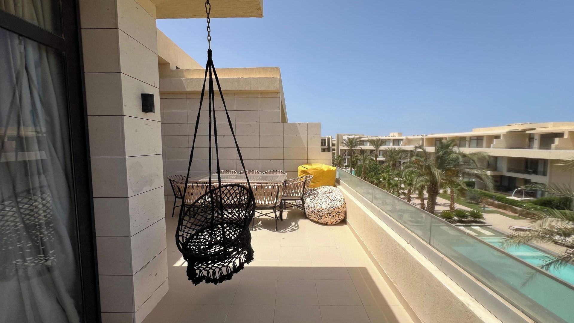 A balcony with a hanging chair, a table, and two ottomans.