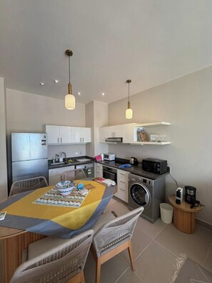 A compact kitchen with a refrigerator, microwave, and washing machine. There is a dining area with a table and chairs. - Cyan Elegant 3BR Golf View Escape (Hurghada)