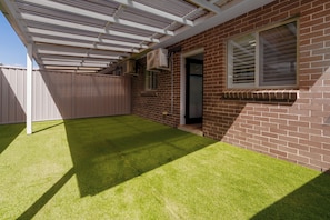 A backyard with artificial grass, a brick wall, a window with blinds, and an air conditioning unit. - A Dahlia by Esteem Retreats (Quakers Hill)