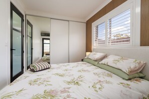 A bedroom with a bed, pillows, a window with blinds, and a door with a mirror. - A Dahlia by Esteem Retreats (Quakers Hill)