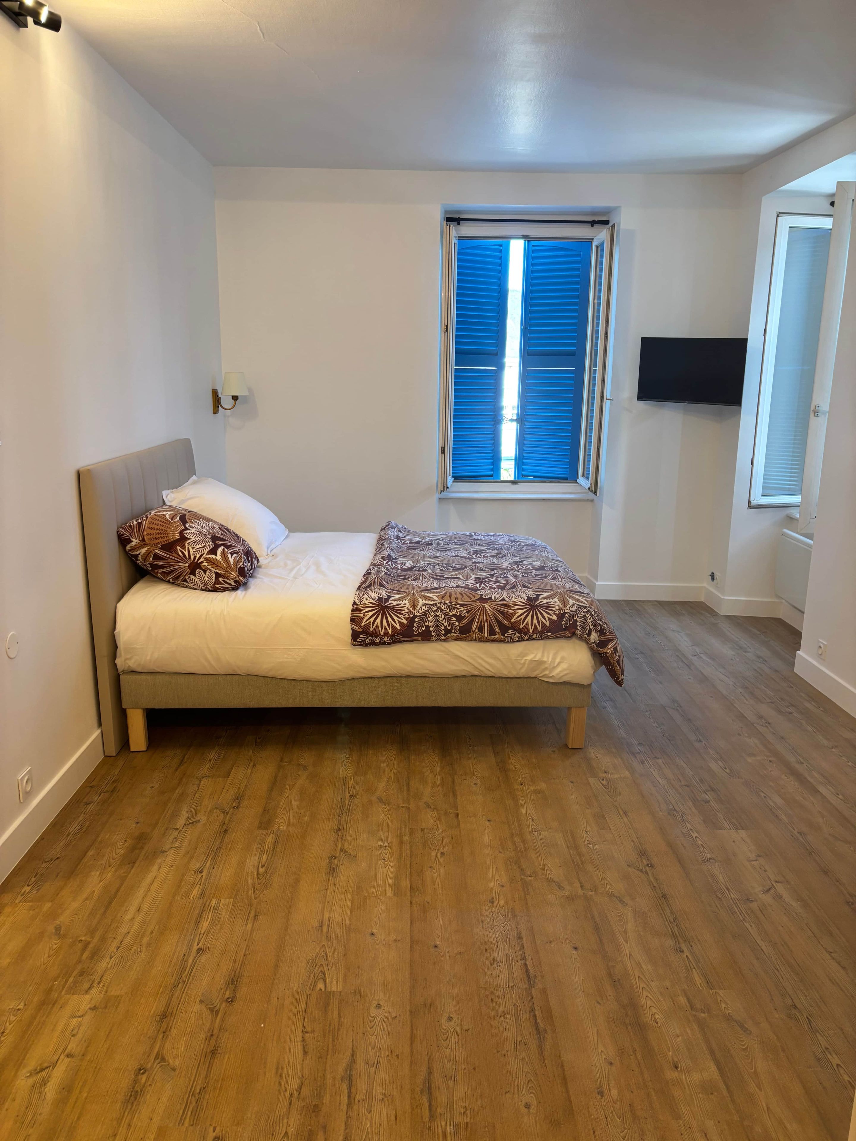 Een slaapkamer met een bed, een raam met jaloezieën, een aan de muur bevestigde televisie en een deur.