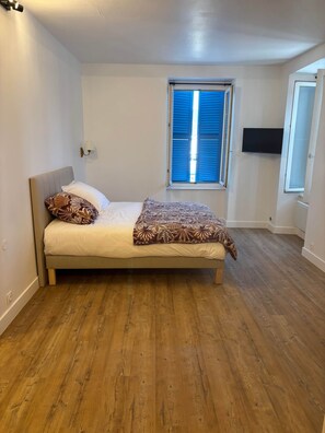 A bedroom with a bed, a window with blinds, a wall-mounted TV, and a door.