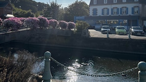 A canal with a bridge, a row of parked cars, and buildings with signage in a sunny setting. - TYYANN 9: A 30-square-meter studio equipped with a kitchenette and a bathroom (Plancoët)