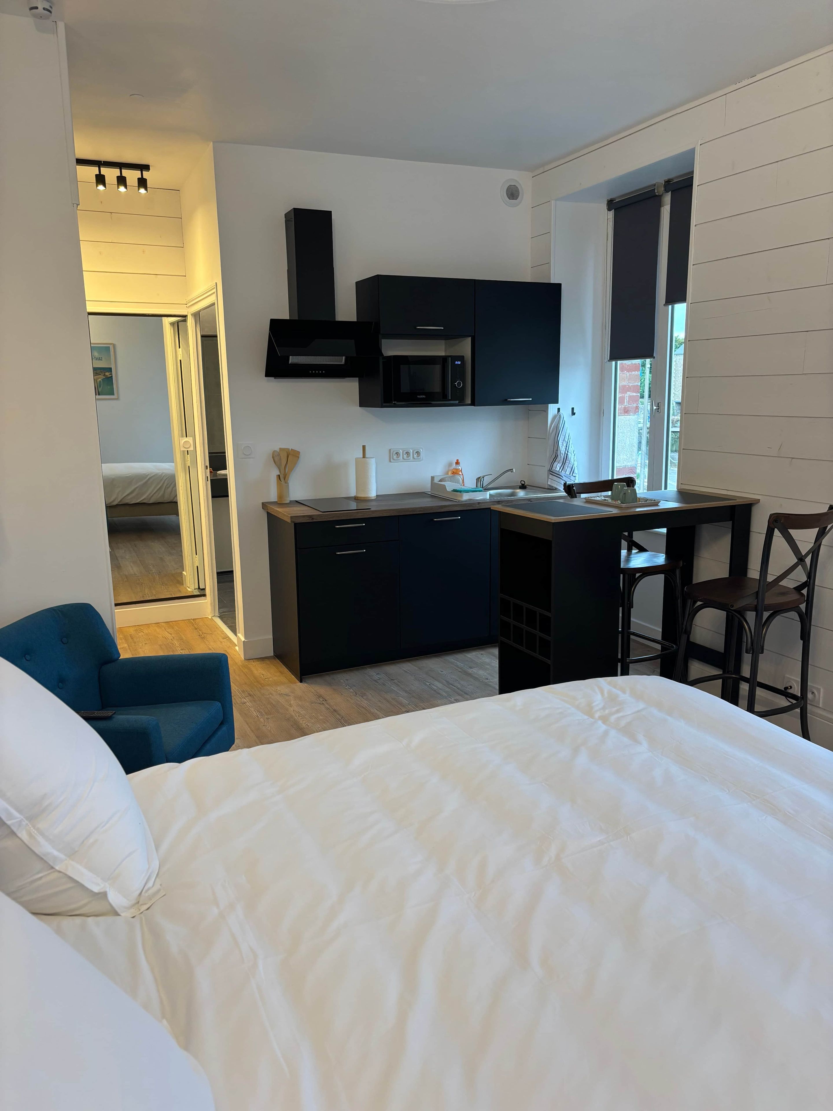 A modern bedroom with a large bed, a desk with a chair, a kitchenette, and a bathroom visible through an open door.