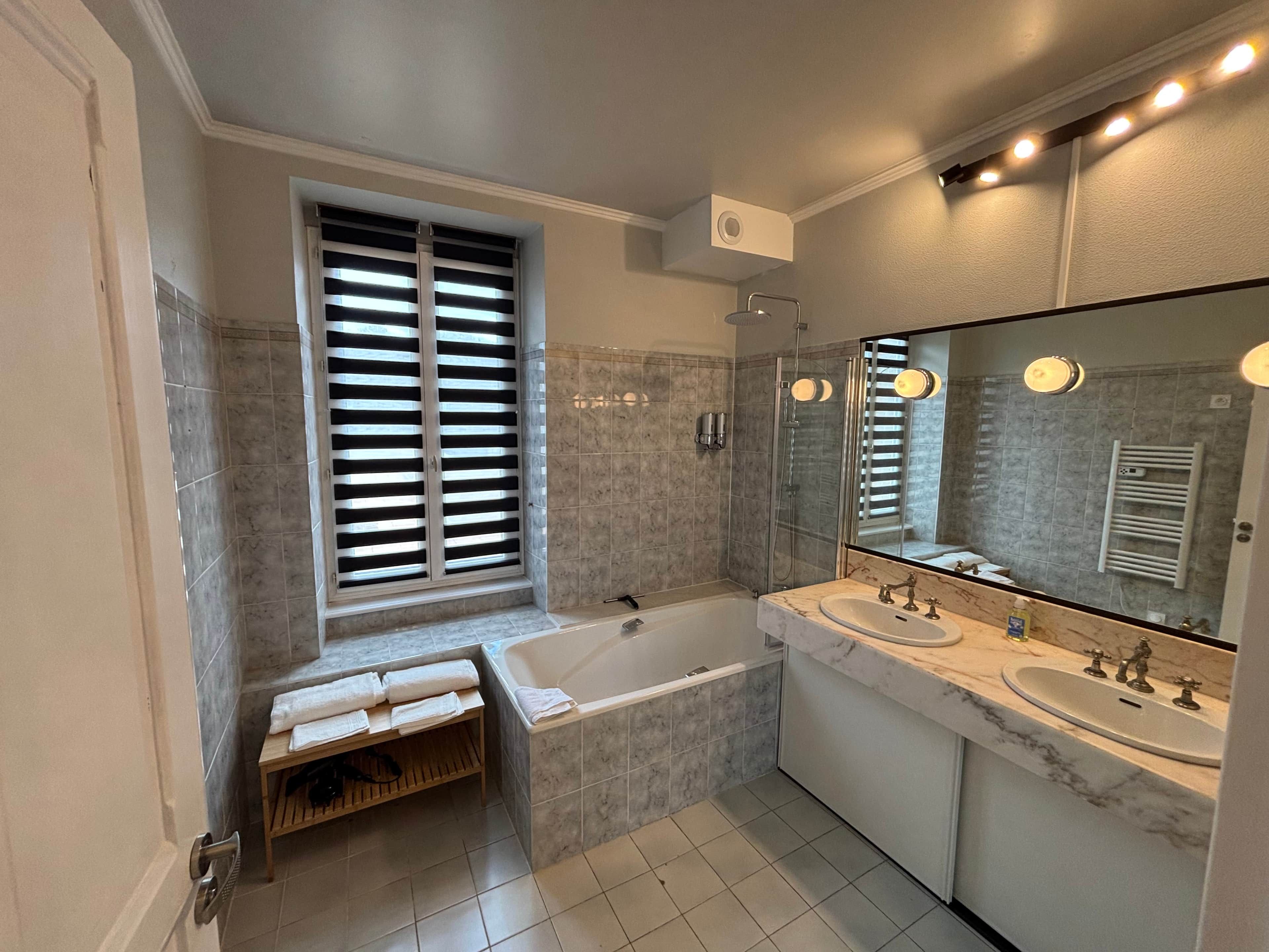Une salle de bain moderne avec une baignoire, deux lavabos, un grand miroir et une fenêtre avec des stores.
