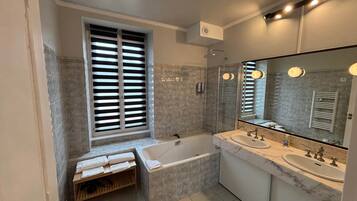 Une salle de bain moderne avec une baignoire, deux lavabos, un grand miroir et une fenêtre avec des stores.