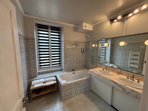 Une salle de bain moderne avec une baignoire, deux lavabos, un grand miroir et une fenêtre avec des stores.