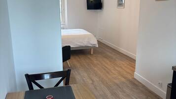 Une chambre de taille modeste, équipée d’un lit, d’un téléviseur fixé au mur et d’un coin repas avec une table et des chaises.