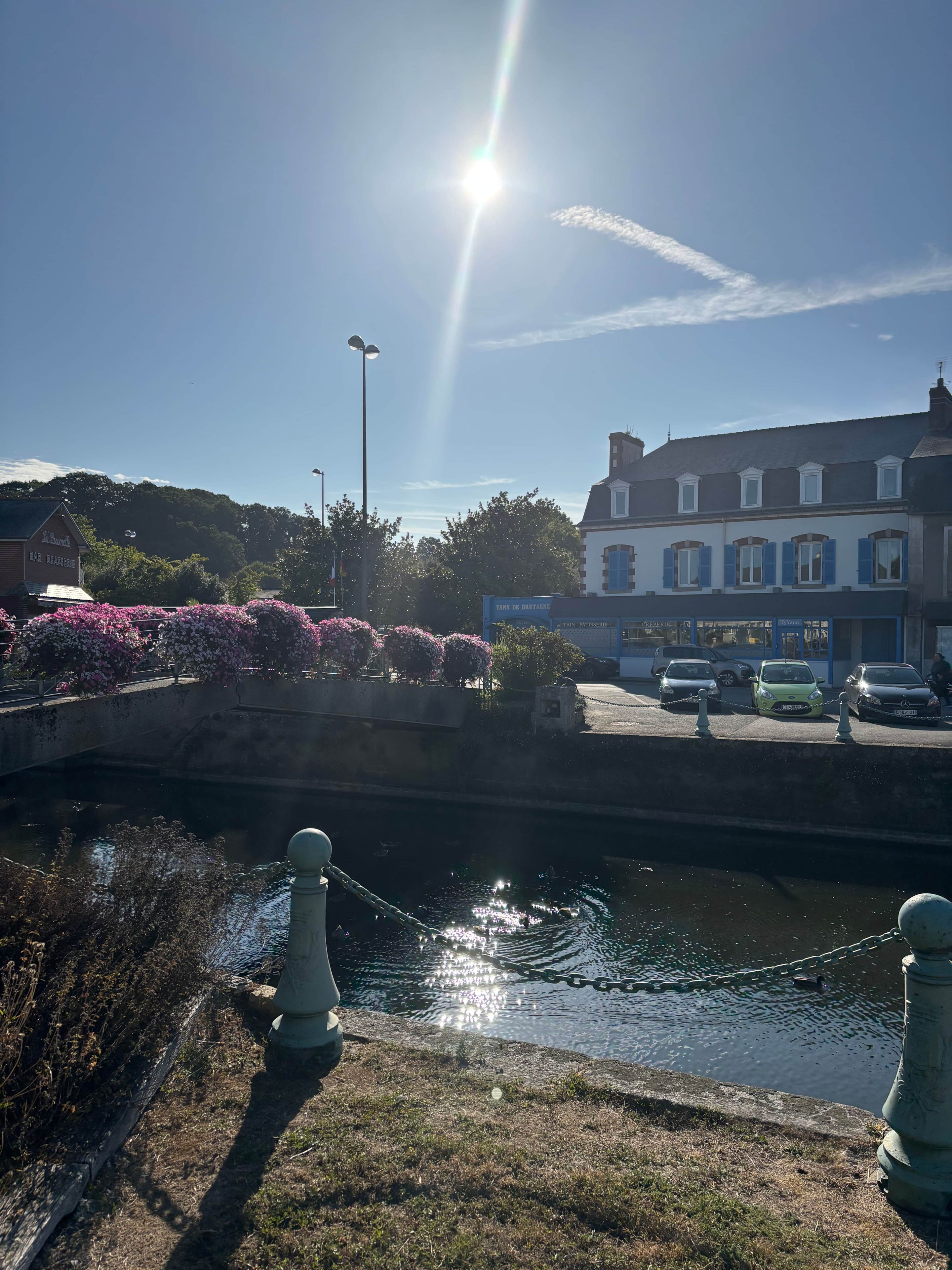 Un canal surmonté d’un pont, une rangée de voitures garées et des bâtiments avec des enseignes, le tout baigné de soleil.
