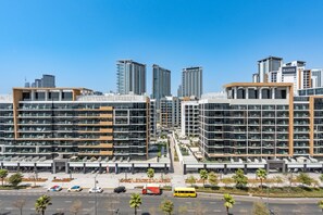 A modern multi-story hotel with a central atrium, multiple balconies, and a parking area with a bus stop. - Studio w/ Community View,Near Meydan Golf Club, Approx 15Min Drive to Downtown (Dubai)