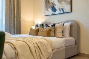 A bed with a beige blanket, multiple pillows, and a wall-mounted abstract painting. - Studio w/ Community View,Near Meydan Golf Club, Approx 15Min Drive to Downtown (Dubai)
