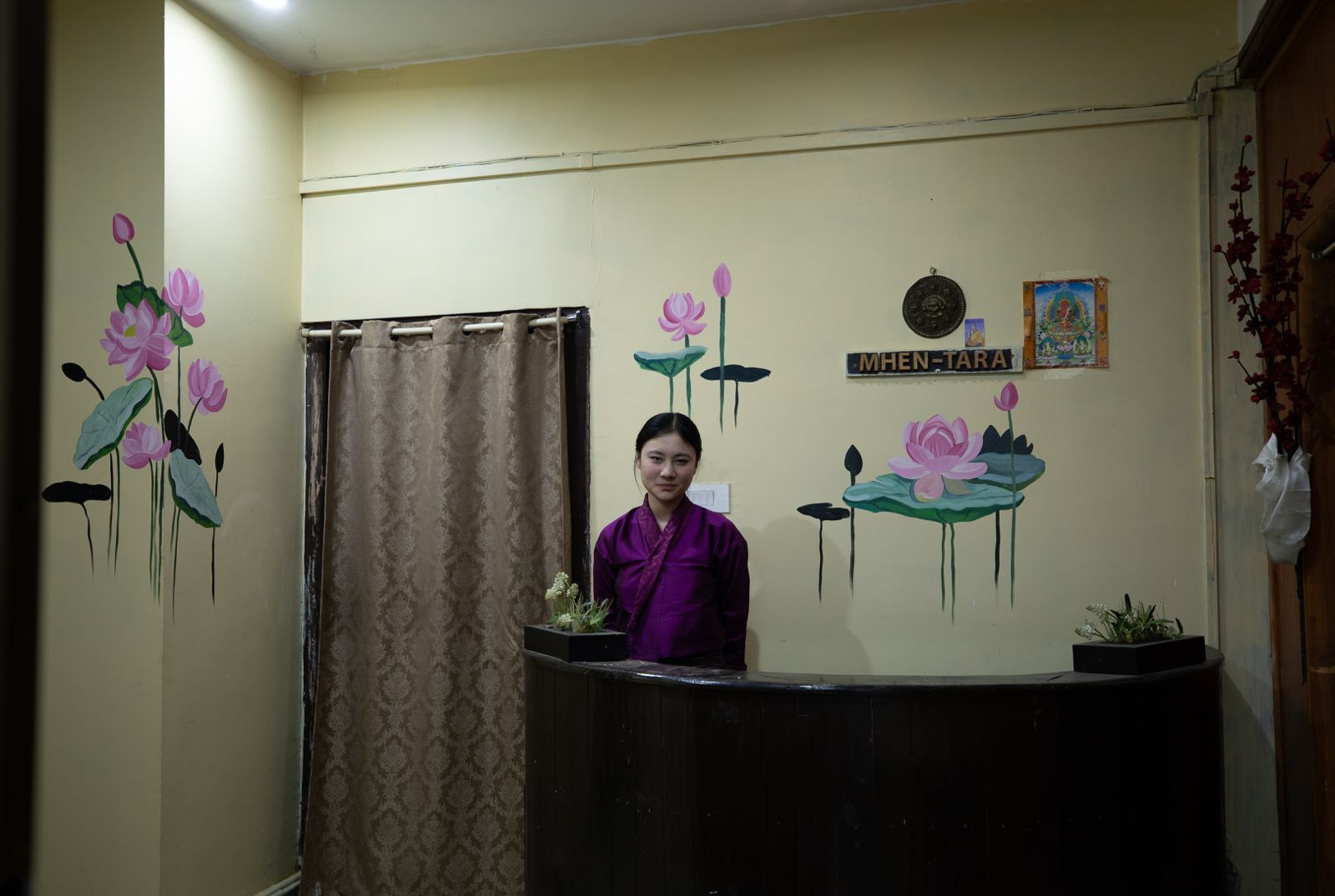 A reception area with a person standing behind a counter, a sign reading 'M-HEN-TARA', and wall art depicting lotus flowers.